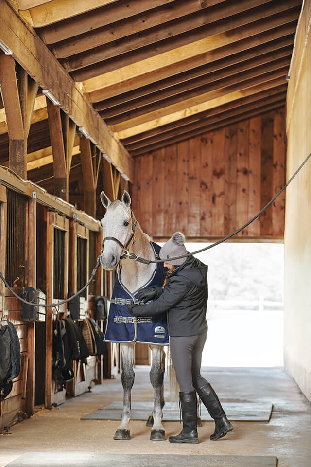 Woman putting a navy turnout on grey horse standing on crossties in barn aisle